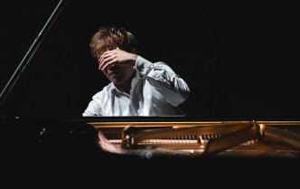 pianista ukazany od przodu, od klatki piersiowej w górę między klapą fortepianu a jego pudłem rezonansowym, uniesioną dłonią zasłania twarz