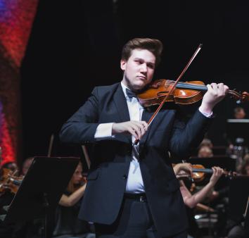 Młody mężczyzna ciemny blondyn gra na skrzypcach w garniturze, za nim orkiestra