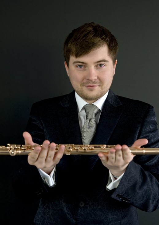 a young man in a dark jacket, visible from the waist up, holding a transverse flute in his hands