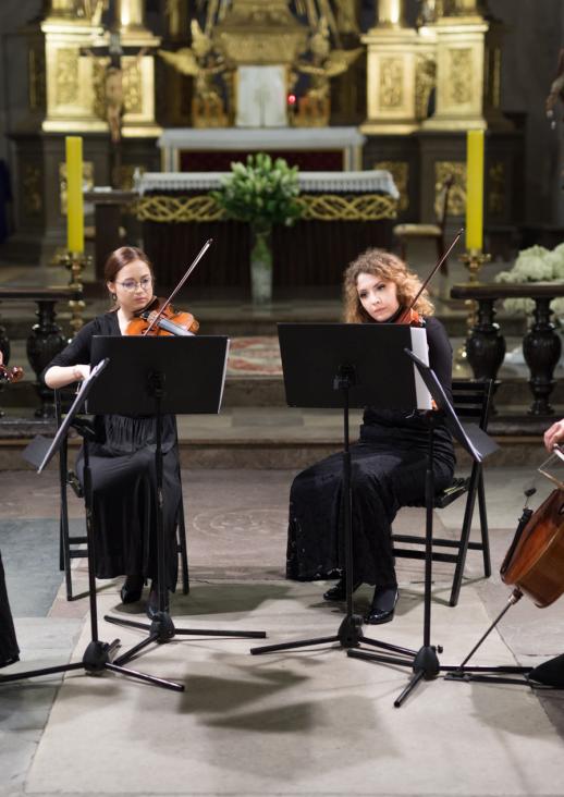 cztery młode kobiety w ciemnych sukniach grające na instrumentach smyczkowych siedzące przed pulpitami