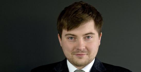 a young man in a dark jacket, visible from the waist up, holding a transverse flute in his hands