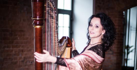 a woman with dark curly hair with a scarf over her shoulders standing with a harp