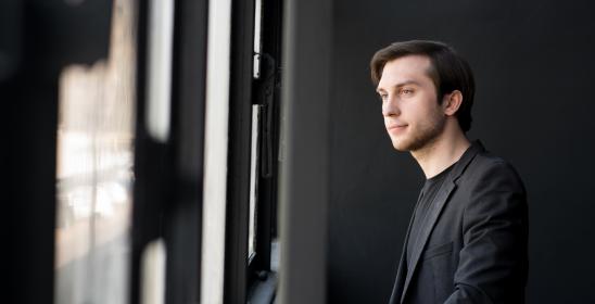 Młody mężczyzna o ciemnych włosach, w czarnej koszuli patrzący przez okno