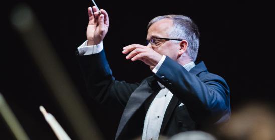 mężczyzna trzymający uniesione ręce, w jednej dłoni trzyma batutę