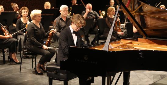 a young man with blonde hair playing the piano, behind him are women and a man with a violin in his hands