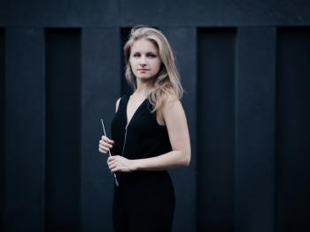 a blonde woman in a dark blouse standing against a dark wall holding a baton in her hands