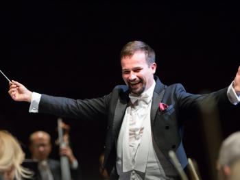 mężczyzna we fraku z rozłożonymi rękoma i batutą w prawej dłoni 