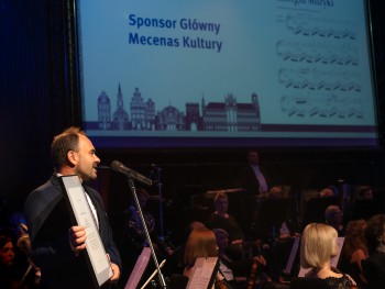 mężczyzna przemawiający przed koncertem, w tle siedząca orkiestra