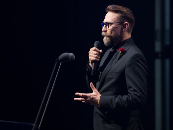 mężczyzna w czarnym garniturze stojący na scenie, trzymający mikrofon w ręce