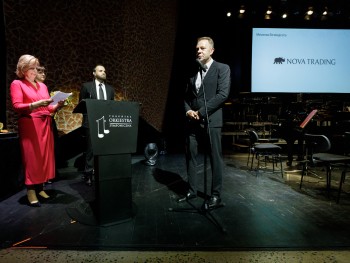 Mężczyzna przemawiający do mikrofonu na scenie, w tle logotyp Nova Trading SA oraz mężczyzna i dwie kobiety na scenie. 