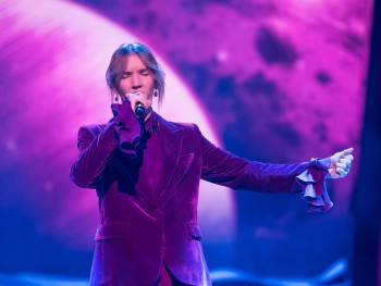 mężczyzna w fioletowym garniturze z żabotami śpiewa do mikrofonu, lewa ręka wyciągnięta jest do boku